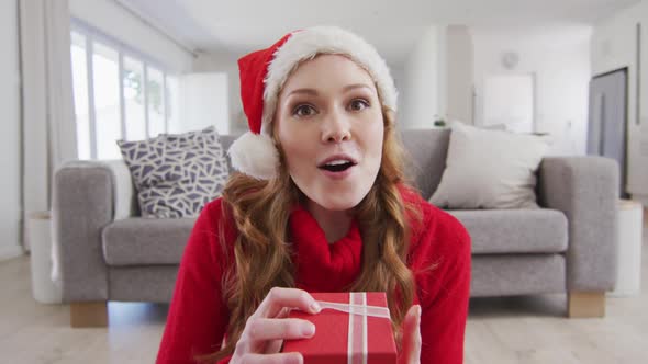 Surprised woman in Santa hat opening a gift box while having a video call on laptop at home