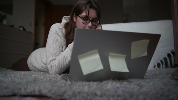 Beautiful girl with glasses in a beige sweater lying on a carpet and watches a movie on a laptop