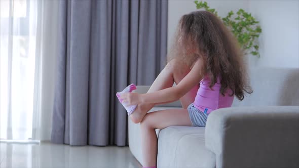 Curious Cute Girl of Preschool Age Sitting on a Sofa Puts White and Pink Socks on Her Legs. Skill