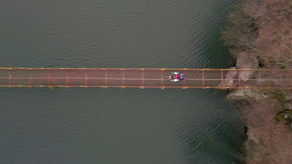 Aerial Slowmotion Shot of Young Sport Man Rides Bicycle on Suspension Bridge Over Small River at