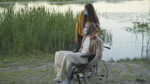 Side View of Caucasian Woman in Face Mask and Disabled Man on Wheelchair Resting on River Bank on