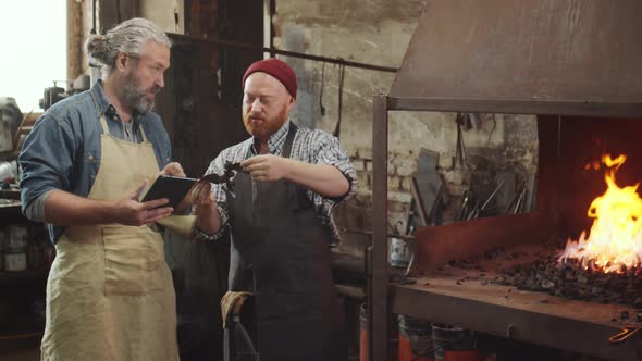 Two Blacksmiths Using Tablet while Working in Smithy