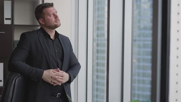 View of a Young Successful Businessman Standing in an Office and Looking Through a Fulllength Window