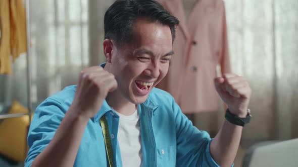 Close Up Of Happy Asian Male Designer Working On A Laptop And Celebrating While Designing Clothes