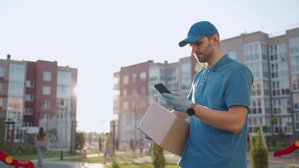 The Postman with Glasses Carries the Parcel and Looks at the Delivery Address Via Mobile Phone