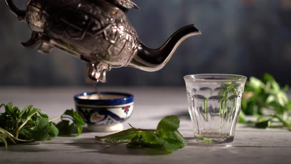 Wide shot of hand picking up an ornate teapot and pours tea into a glass with Moroccan bowl and mint