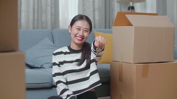 Woman With Cardboard Boxes Sit On The Floor Smiling And Showing The Keys To Camera In The New House