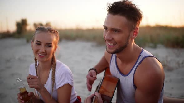 Beautiful Couple Smiling Resting Playing Guitar at Coast at Sunset Slow Motion