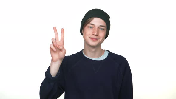 Portrait of Smiling Caucasian Youngster Wearing Hat Showing Two Fingers Victory Sign Over White