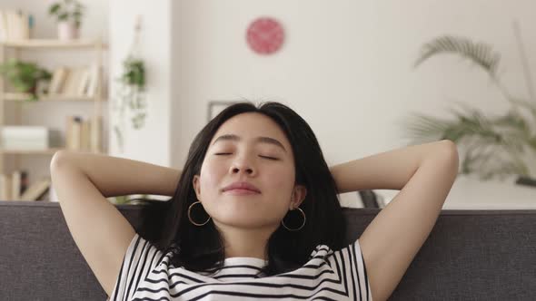 Young Asian Woman with Hands Behind Head Relaxing on Sofa at Home