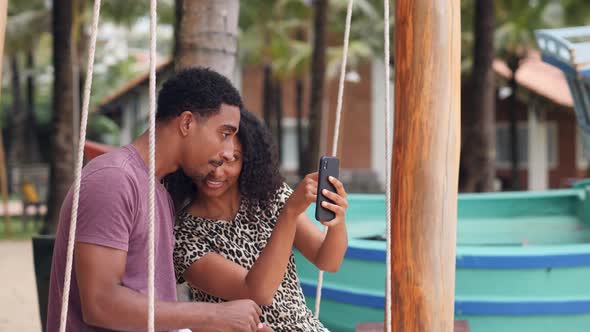 Happy Couple Sitting on a Swing Taking Selfie Photos Together By Smartphone