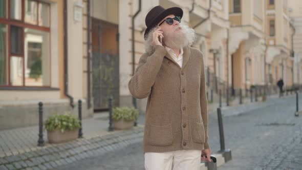 Portrait of Cheerful Old Man with Long Grey Hair and Beard Talking on the Phone While Walking with