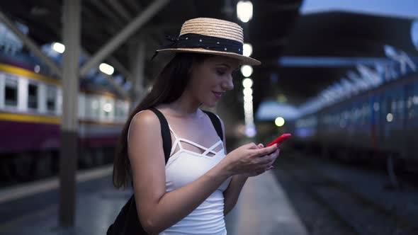 Young Brunette Woman Traveler Laughs While Playing a Mobile Phone in Train Station Platform