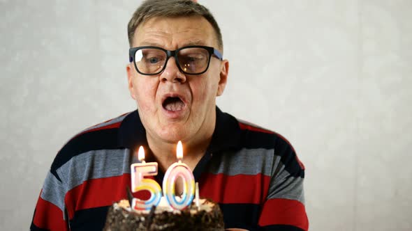 Joyful Mature Adult Man Wearing Blue Eyeglasses Blows Out Number 50 Candles on Birthday Cake.