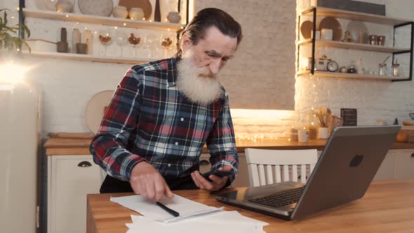 Elderly Man Working on Laptop and Writing Notes in Notebook Sitting at Table