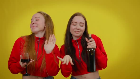 Positive Twin Sisters Dancing with Wine Bottle and Glass of Drink at Yellow Background