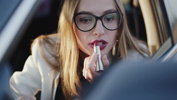 Beautiful Woman Looking in Rear View Mirror and Applying Red Lipstick in Car