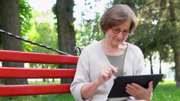 Senior Woman of Retirement Age Sitting on Park Bench Making Video Call Looking at Tablet Camera