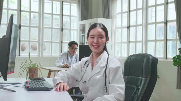 Young Asian Woman Doctor Is Using Computer, She Turns And Smiles To Camera In Workplace