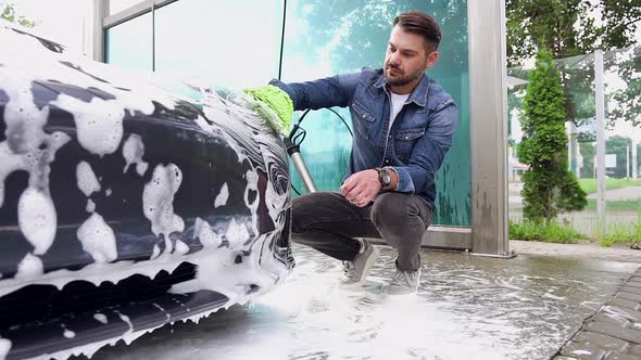 Bearded Guy which Squatting Near His Trendy Car and Washing Headlights with Foamy Sponge in Car Wash