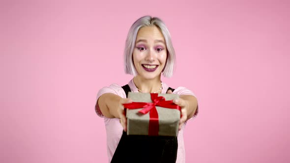 Cute Young Woman with Colorful Hair Holding Gift Box with Bow and Gives It By Hands To Camera on
