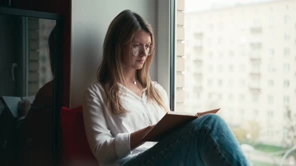 A Young Woman with Glasses Sits on the Windowsill and Reads a Book.