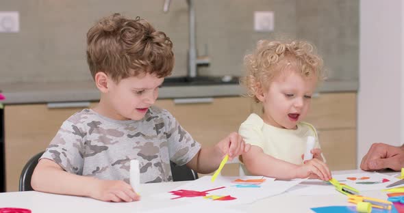 Small Girl and a Small Boy are Making Applications with the Coloured Paper and Glue and Their Father