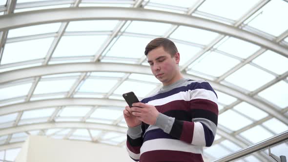 A Young Man Is Standing with a Phone. A Man Uses a Smartphone Against a Pan-glass Roof