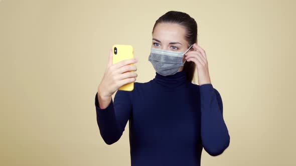 Woman in Medical Mask Makes Selfie on Mobile Phone, Isolated on Beige Background