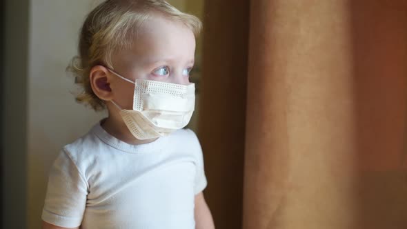 little girl in a protective medical mask stands near the window curtains.