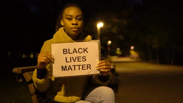 A Young Black Woman Shows a Black Lives Matter Sign to the Camera in a City Park at Night