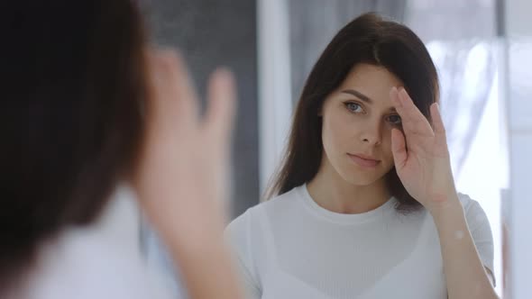 Serious young brunette woman in white shirt looking at herself in the mirror.