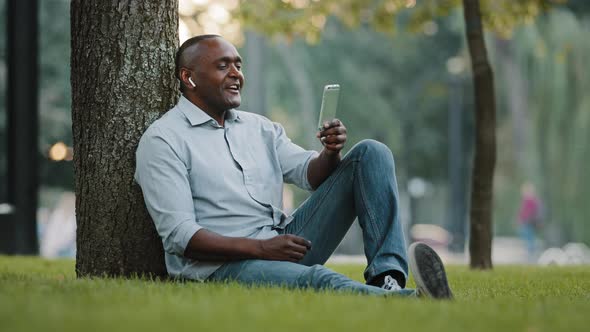 Elderly African Man Entrepreneur Sitting on Grass in City Park Businessman Holding Smartphone