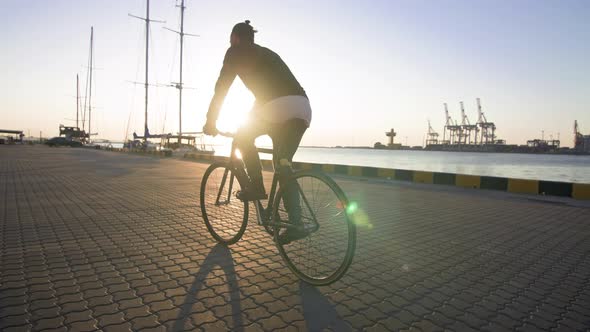 Stylish Handsome Man Riding Bicycle During Sunset or Sunrise with Sea Port on Background