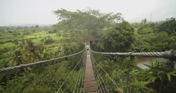 Wide View of a Man Walking Across a Shaky Rope Hanging Bridge Across a Tropical Jungle in Bali in