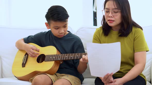 Asian mother embraces son, Asian boy playing guitar and mother embrace on the sofa