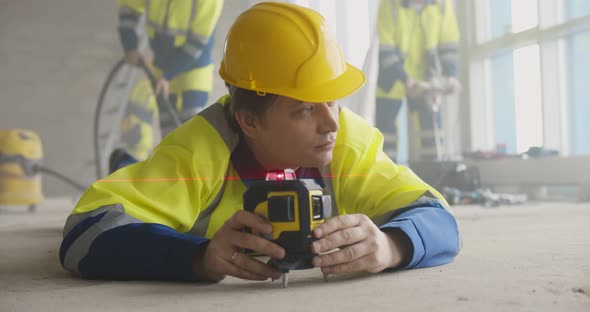 Builder in Uniform Lying on Floor Using Laser Level at Construction Site