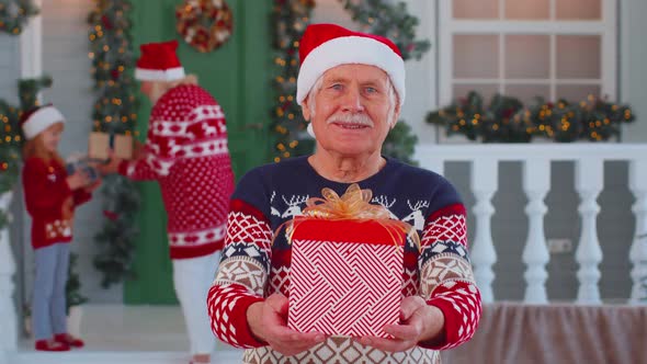 Portrait of Grandfather Man Presenting Gift Box Smiling Near Decorated Christmas House with Family