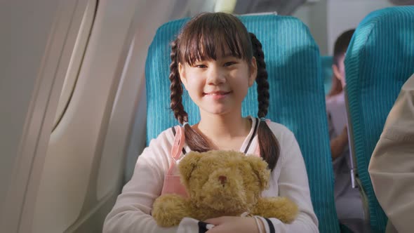 Asian little cute kid holding bear doll, sit on board next to window in airplane and look at camera.