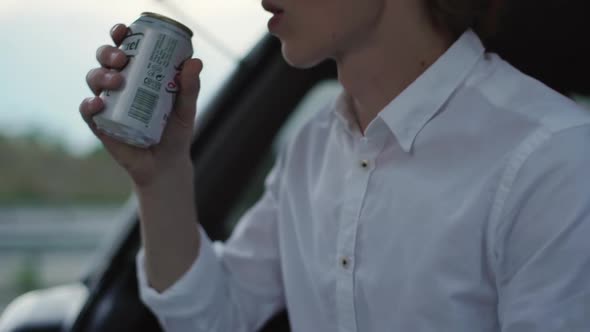 Caucasian Guy in Good Mood Making Pause Sitting in Car on Road During Sunset Drinking Beer From Can