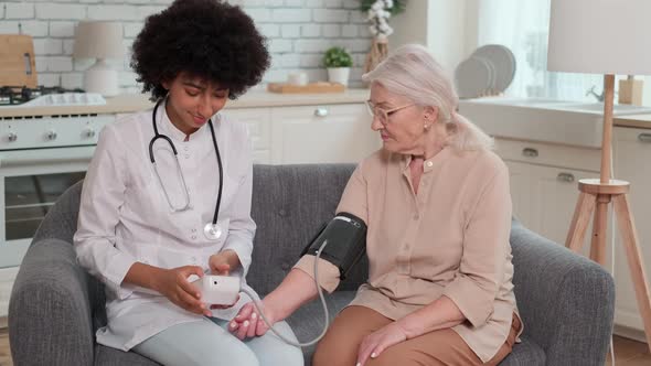 African American Woman Doctor Checking Blood Pressure of Senior Woman While Sitting on Couch at Home