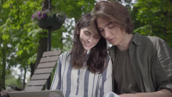 The Male and Female Students in Casual Clothes Sitting at the Bench, Girl Holding Laptop and Boy Has