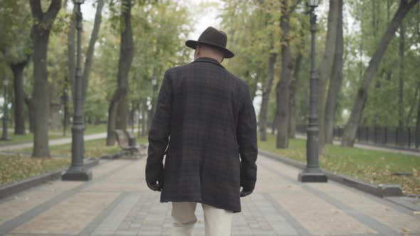 Back View of Elegant Man in Coat, Hat, and Gloves Walking Along Spring or Autumn Park. Camera