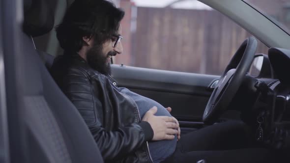 Side View Close-up of Funny Pregnant Man Sitting at Driver's Seat and Caressing His Belly