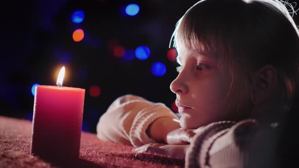 A Girl Looks at a Burning Candle, Sits on the Background of the New Year Tree in the Evening