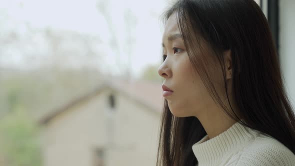 A Young Disappointed Asian Woman is Sitting in the House and Looking Out the Window