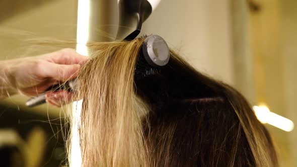 Lady Straightens Blonde Hair of Client with Brush and Dryer