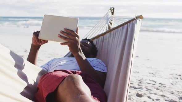 African american man using digital tablet while lying on a hammock at the beach