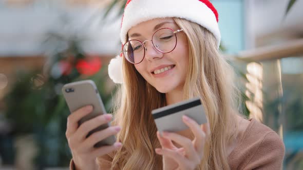 Caucasian Girl in Santa Hat Makes Internet Order Using Telephone and Credit Card Millennial Woman