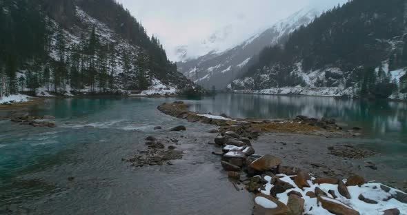 flying between spruces trees towards a stone pier in blue cyan lake in mountains during heavy snow